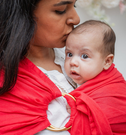 close up of mom and baby in a ring sling carrier. the mom is kissing baby's head. the ring sling has gold rings and is made of linen in a red/pink colour