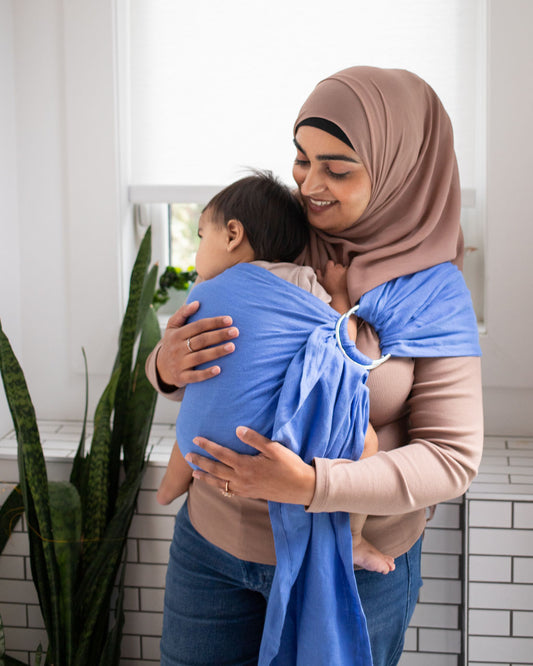 Woman wearing a baby in a blue sling against a white tiled wall.