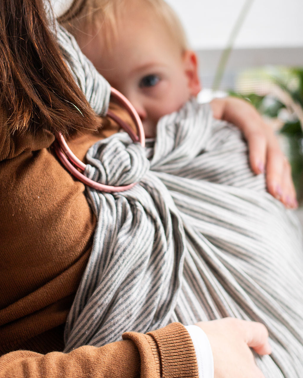 Person holding a baby wrapped in a gray stripe ring sling, with a blurred background