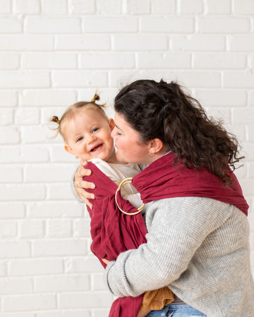 mom kissing toddler wrapped in a red sling against a white brick wall.