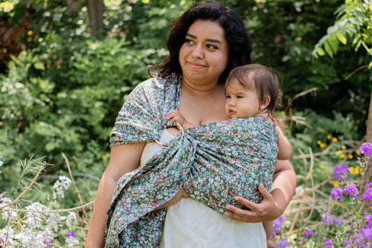 Woman holding a baby wrapped in a floral sling amidst flowers and greenery