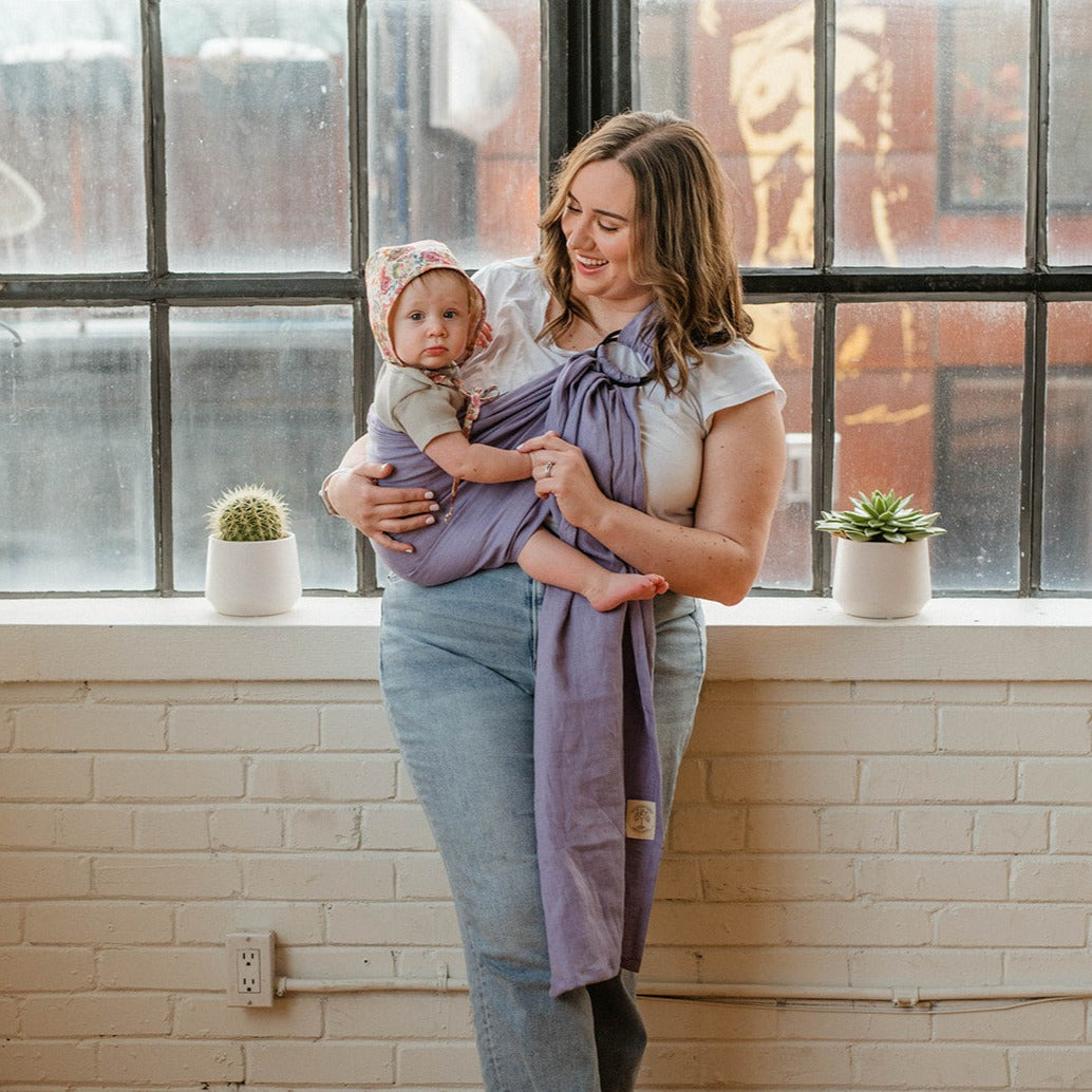 A mom is standing by a window, wearing a purple ring sling baby carrier , holding a baby who is facing the camera.