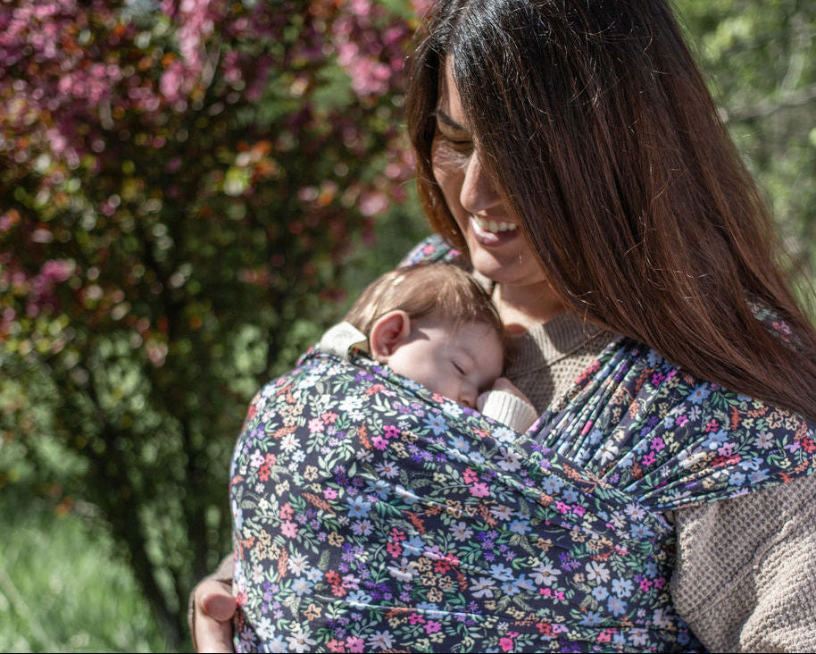 Woman holding a baby in a floral baby carrier outdoors with greenery in the background
