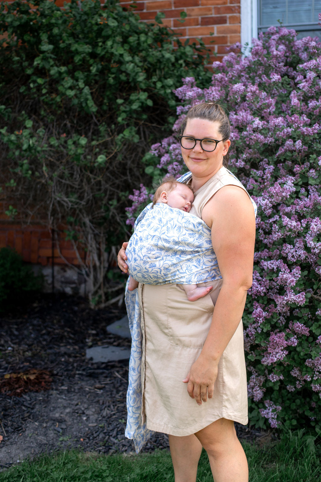 Woman holding a baby wrapped in a ring sling outdoors with greenery and flowers in the background