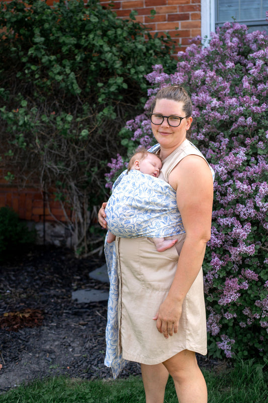 Woman holding a baby wrapped in a ring sling outdoors with greenery and flowers in the background