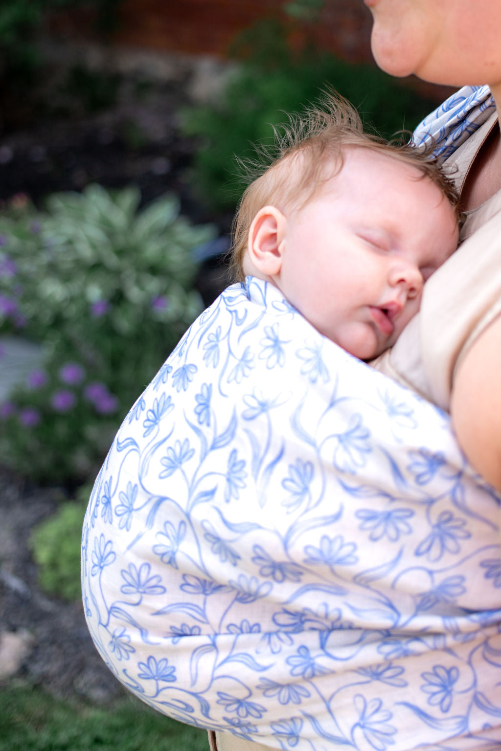 Baby sleeping in a blue and white floral baby carrier with a blurred outdoor background
