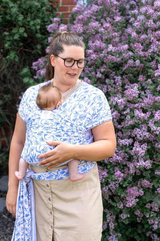Woman holding a baby in a white with blue floral pattern stretchy wrap carrier with purple flowers in the background