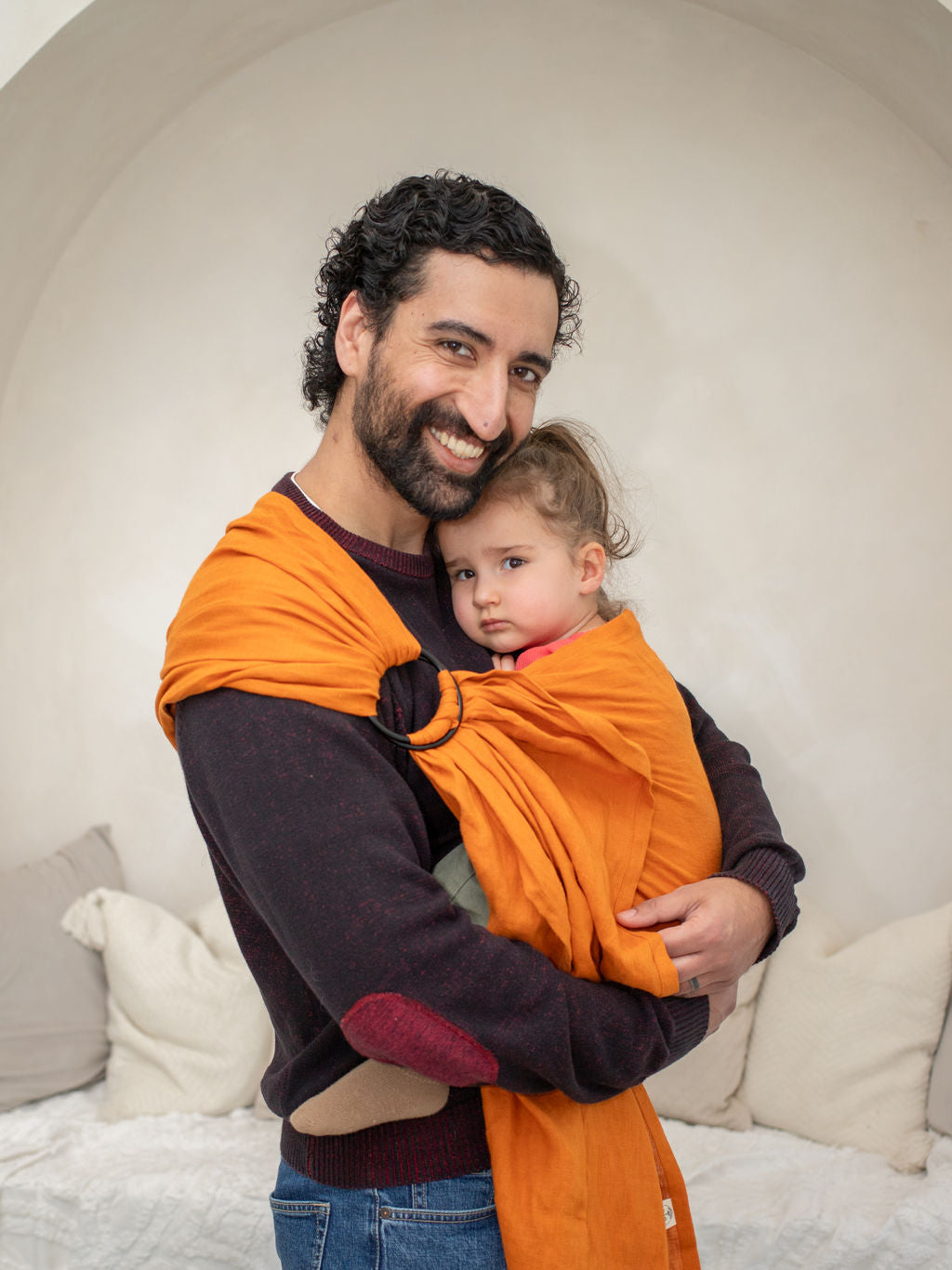 Man holding a toddler in an orange ring sling carrier against a neutral background