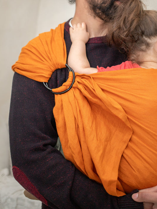 dad wearing an orange ring sling baby carrier with a toddler inside, against a neutral background.