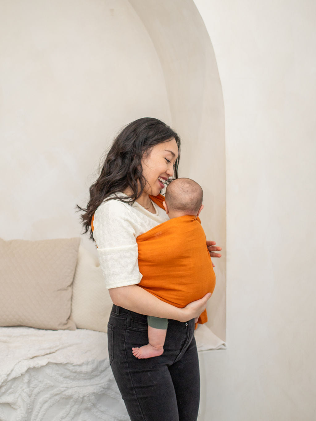 Woman carrying a baby wrapped in an orange sling against a neutral background