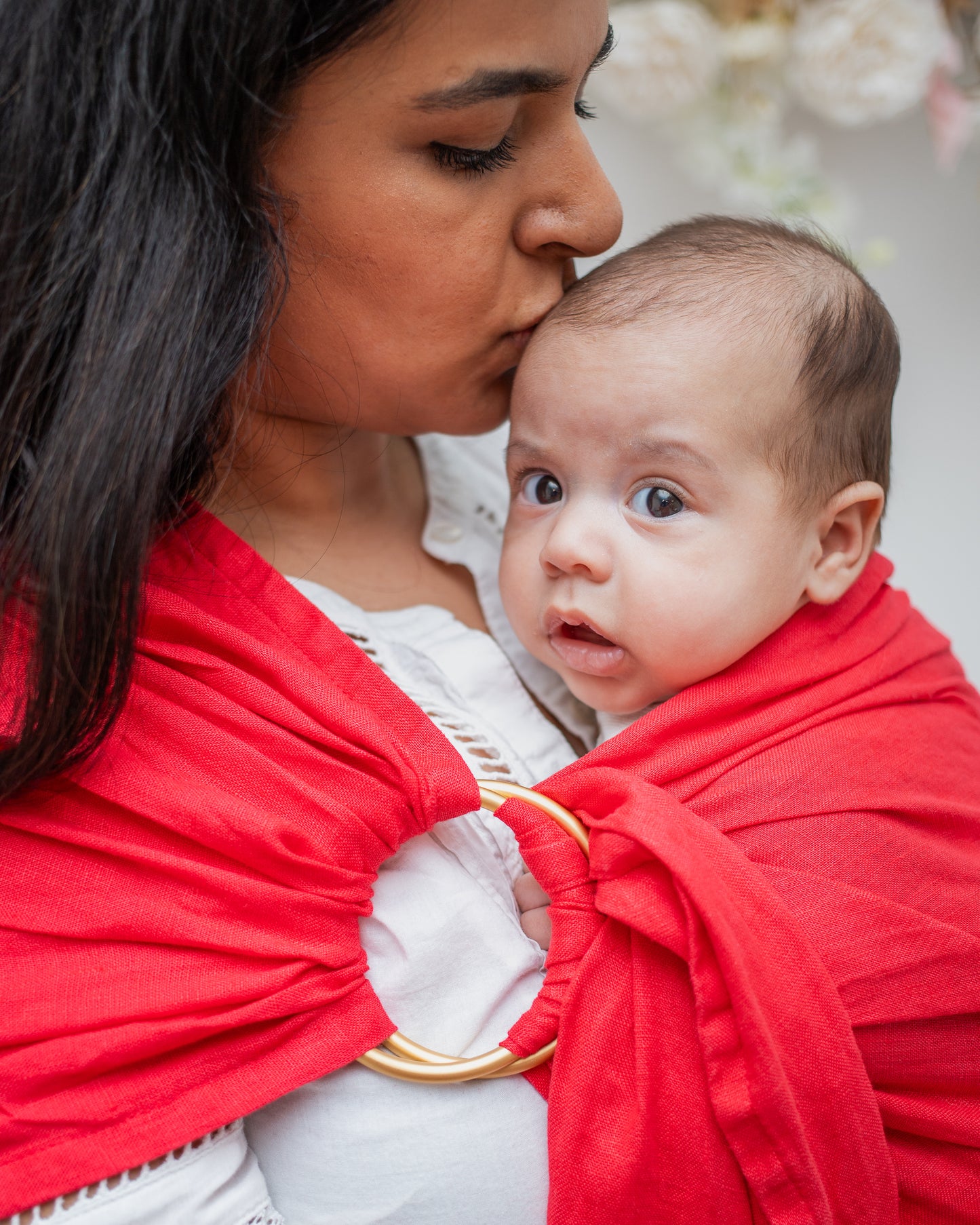 close up of mom and baby in a ring sling carrier. the mom is kissing baby's head. the ring sling has gold rings and is made of linen in a red/pink colour