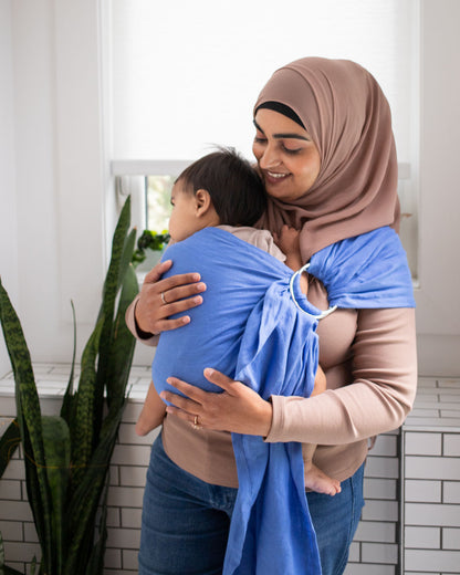 Woman wearing a baby in a blue sling against a white tiled wall.