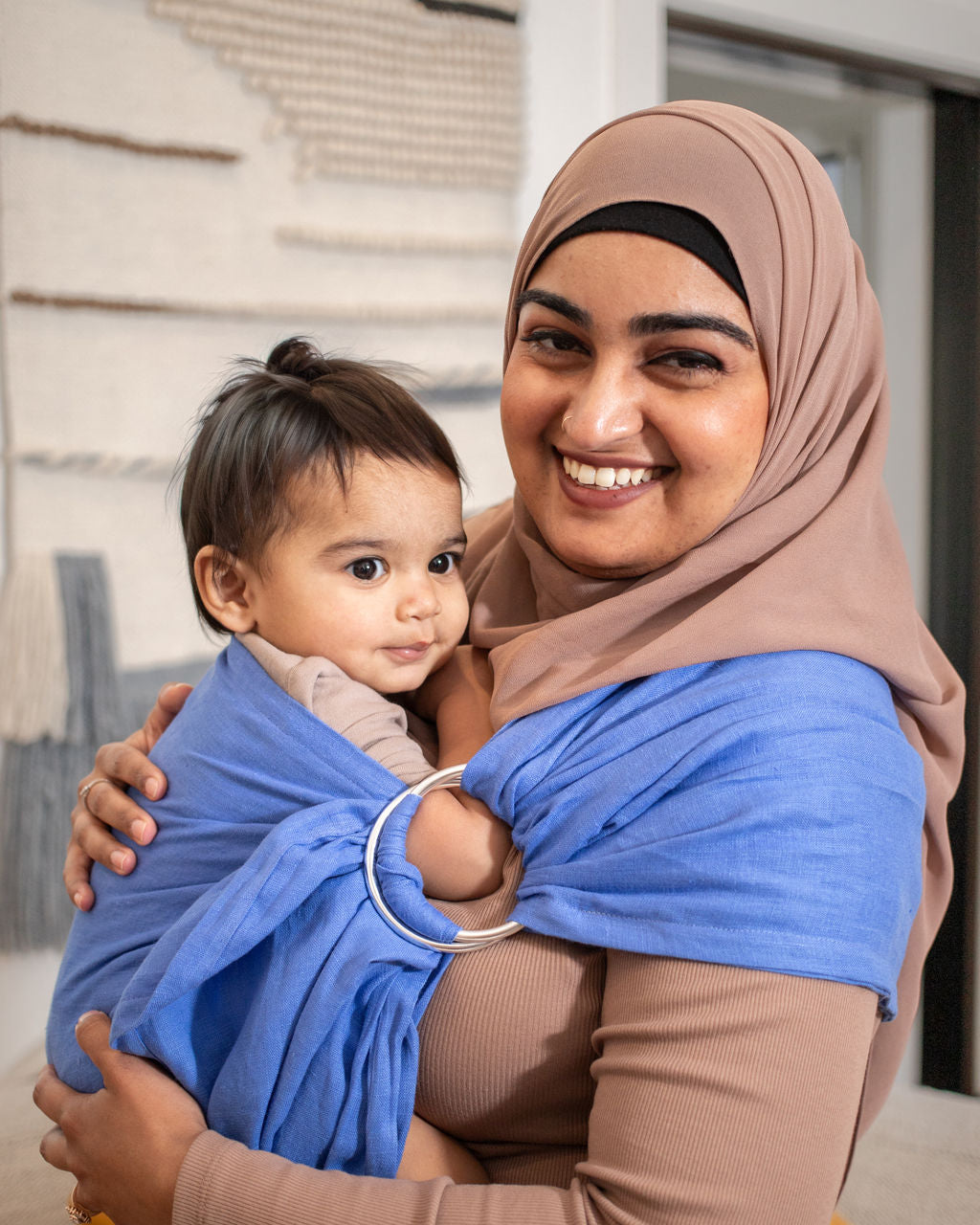 Woman wearing a brown hijab holding a child wrapped in a blue linen ring sling