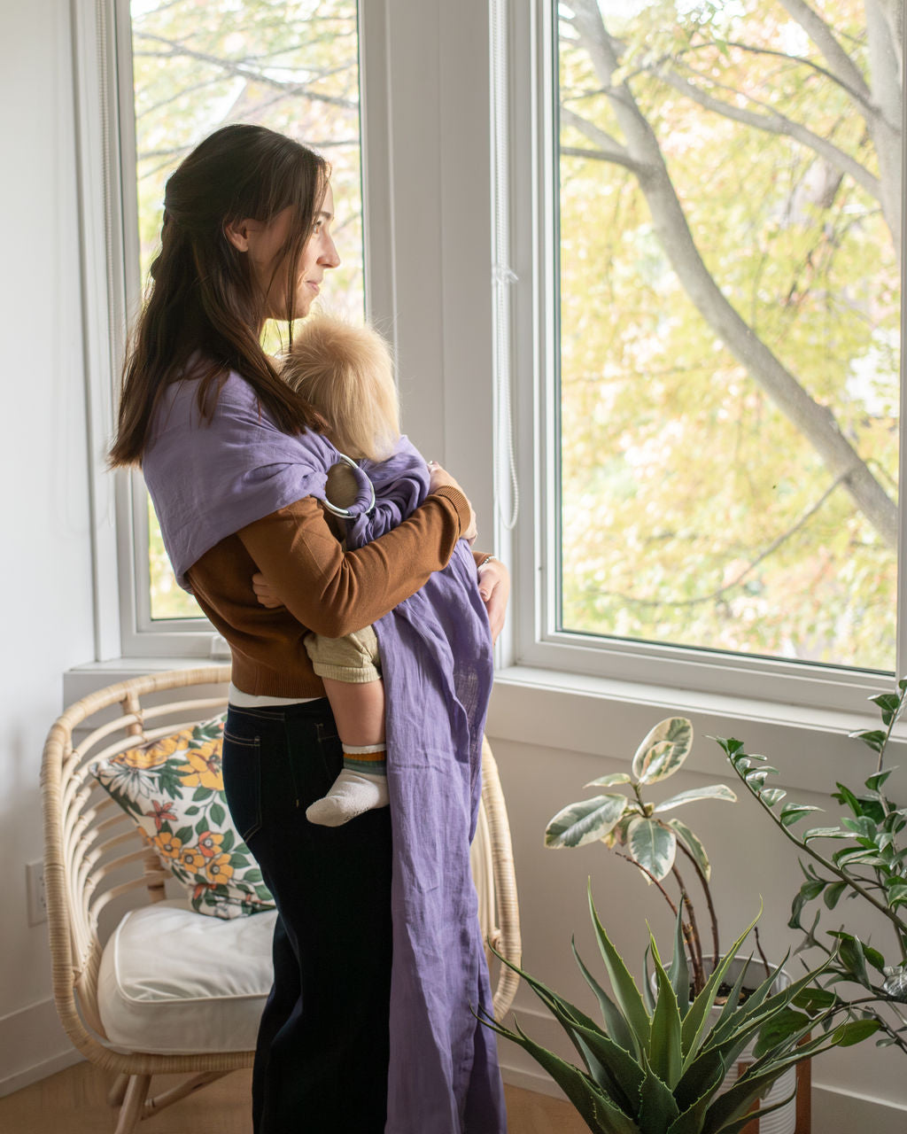 Mom holding a toddler in purple linen ring sling in front of a window with a plant in the foreground