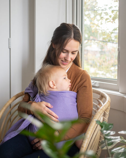Woman holding a baby wrapped in a purple sling indoors near a window.
