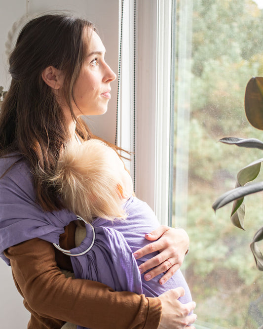 Woman holding a baby in a purple sling by a window