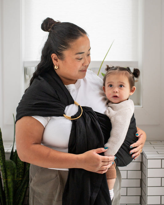 mom holding a child in a black ring sling carrier in a kitchen setting.