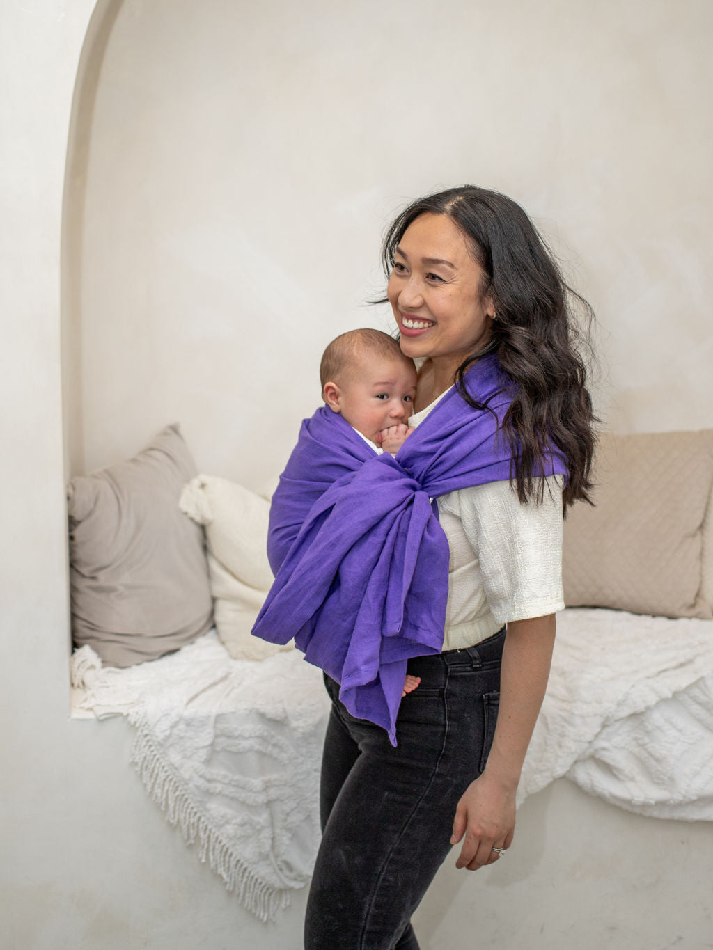 Woman holding a baby wrapped in a purple ring sling baby carrier against a neutral background