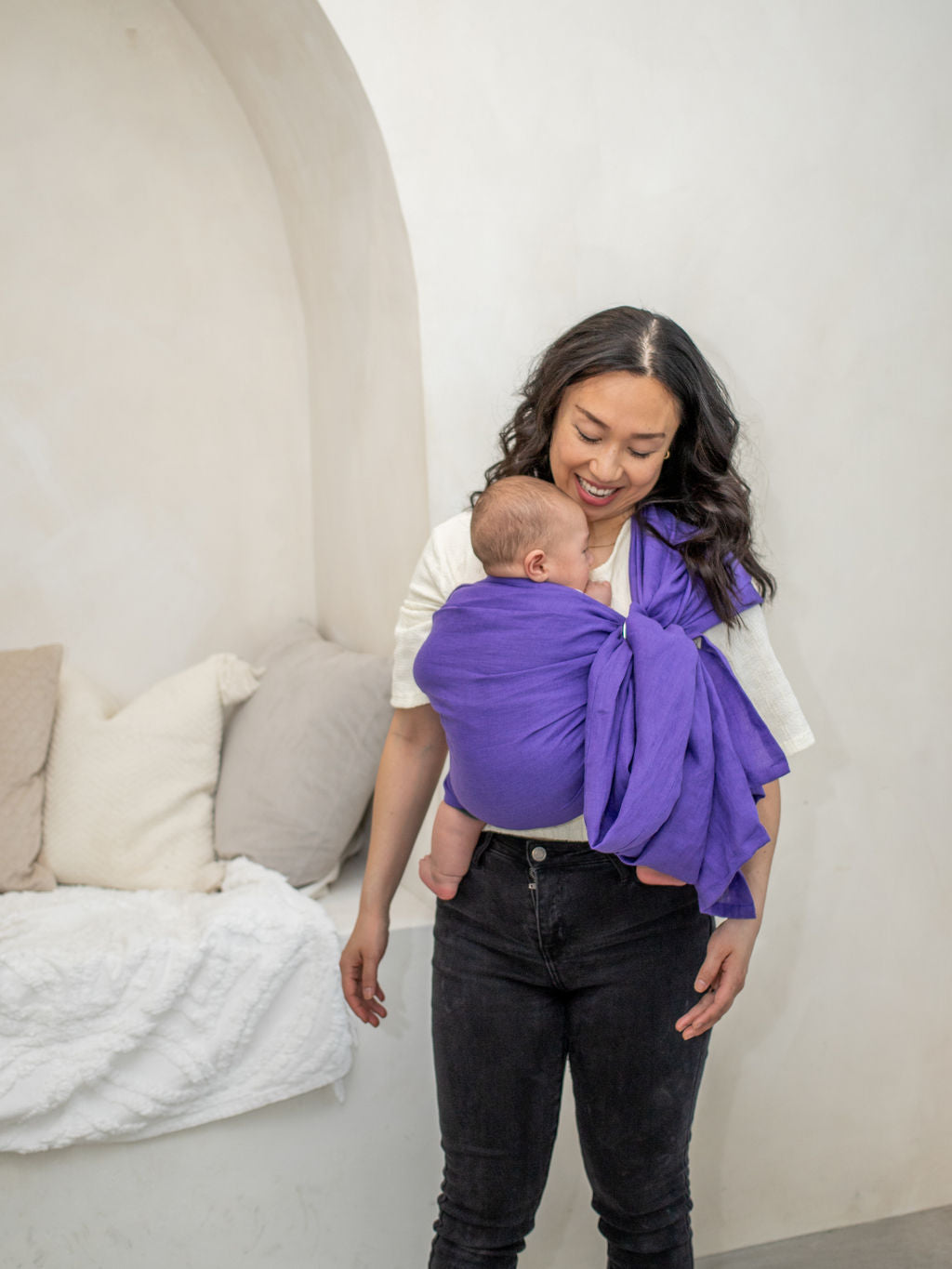 Woman holding a baby in a purple linen sling against a white wall.