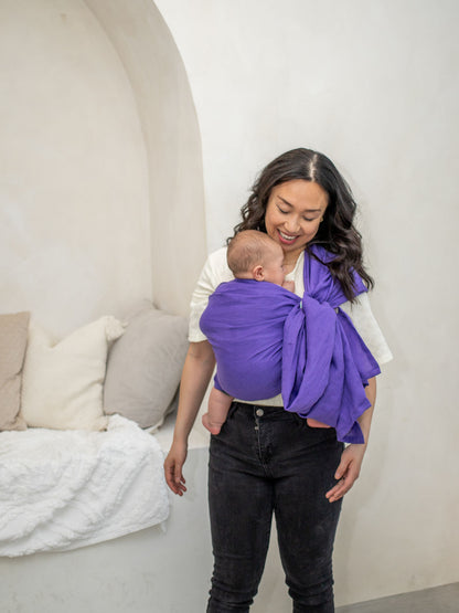 Woman holding a baby in a purple linen sling against a white wall.