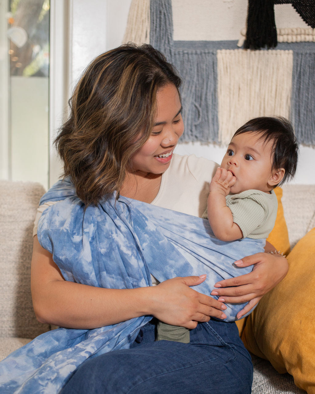 Woman wearing a baby in a hand-dyed sling, sitting on a couch
