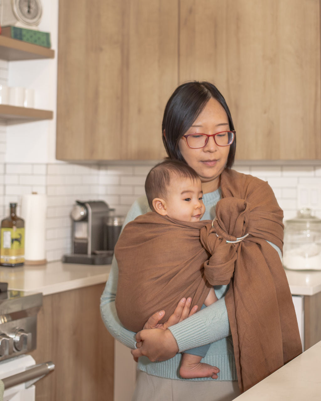 Woman holding a baby wrapped in a brown sling carrier in a kitchen.