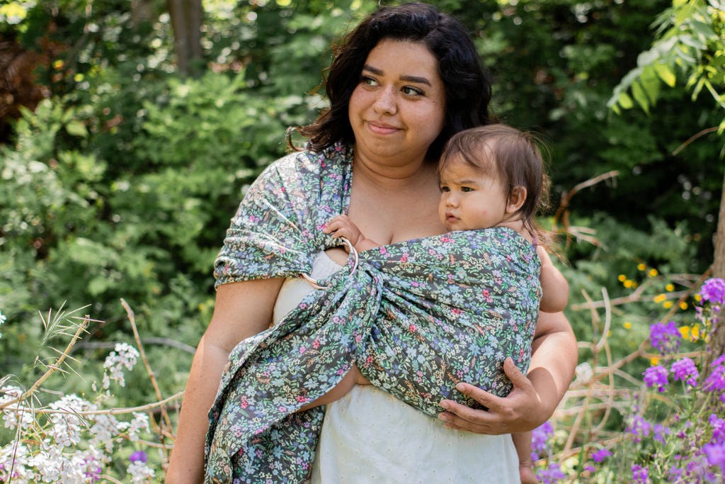 Woman holding a baby wrapped in a floral sling amidst flowers and greenery
